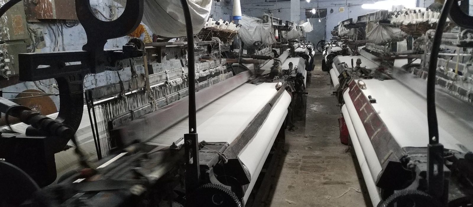 Rows of power looms weaving white cloth inside a textile shed in India
